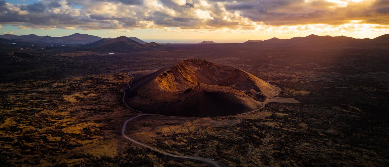 Luftfoto af Vulkanen Cuervo på Lanzarote, med krateret oplyst ved solnedgang og et vulkansk landskab omkring.