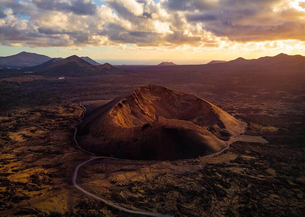 Luftfoto af Vulkanen Cuervo på Lanzarote, med krateret oplyst ved solnedgang og et vulkansk landskab omkring.